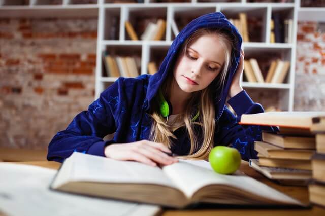 młoda dziewczyna w trakcie back to school siedzi w bibliotece w granatowej bluzie z kapturem na głowie i zielonymi słuchawkami zawieszonymi na szyi czyta książkę na której stoi zielone jabłko