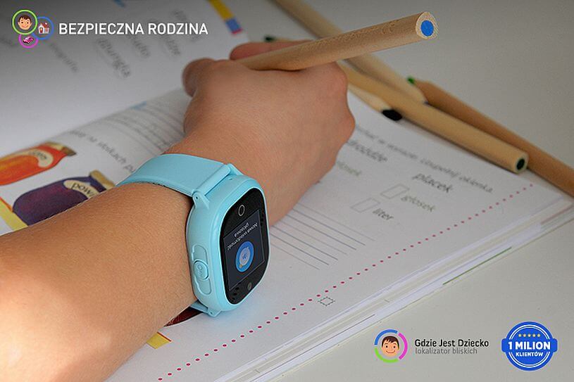 Domowa kwarantanna, sprawdzanie, co robi dziecko, czyli ręka chłopca z niebieskim zegarkiem z GPS od Bezpiecznej Rodziny ułożona na otwartym zeszycie wśród kolorowych kredek