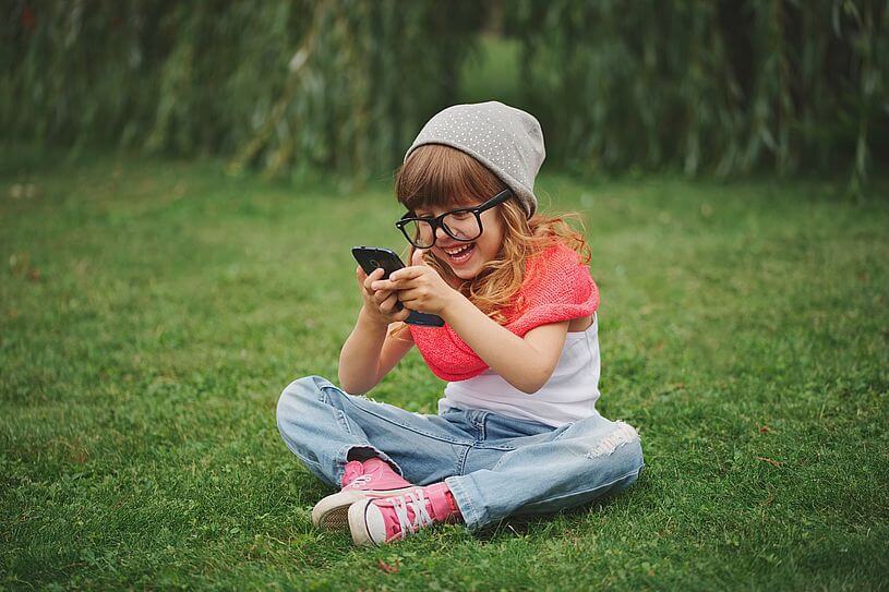 Namierzanie telefonu dziecka, czyli kilkuletnia dziewczynka ubrana w jeansy, białą koszulkę, różowy szalik, różowe trampki i duże czarne okulary, siedzi na trawie, trzymając w wyciągniętych rękach telefon komórkowy i patrzy w niego z uśmiechem