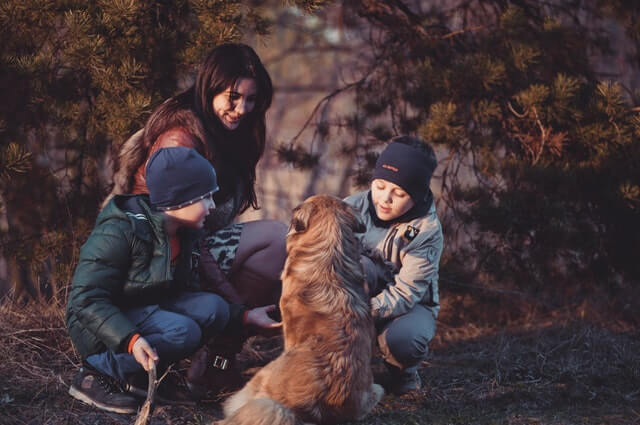 zdjęcie kobiety kucającej przy kucających kilkuletnich chłopcach ubranych w ciepłe kurtki i czapki w lesie przy których siedzi tyłem niewielki pies z beżową sierścią