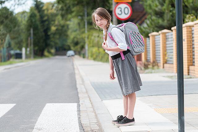 Kilkuletnia dziewczynka z warkoczami na głowie, ubrana w spódniczkę do łydek i białą bluzkę, stoi na przejściu dla pieszych i obserwuje, czy swobodnie może przejść przez ulicę