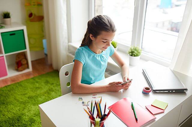 kilkuletnia dziewczynka w ciemnych włosach, ubrana w niebieską koszulkę, siedzi w swoim pokoju przy białym biurku, na którym leży laptop, notes i pojemnik z kredkami i pisze coś na telefonie komórkowym