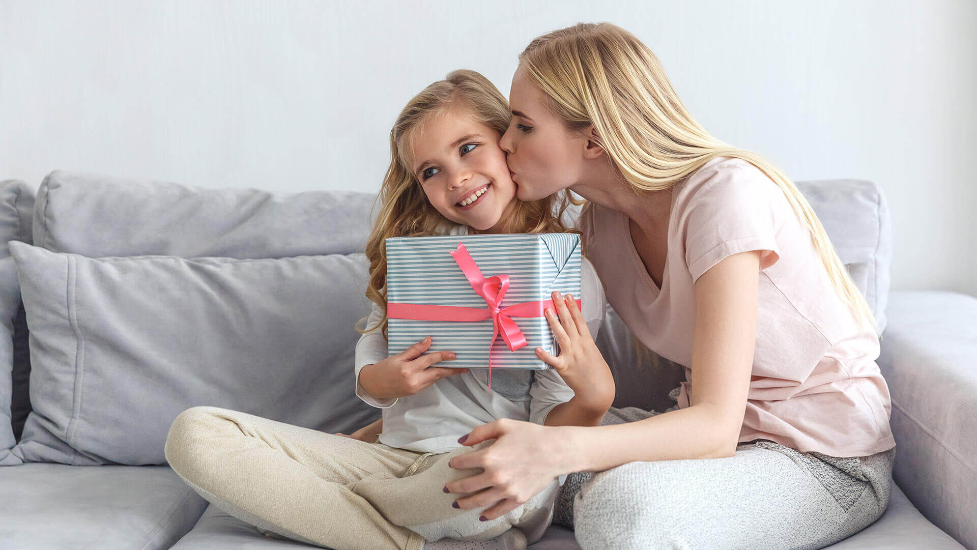 Młoda mama siedząca z córką na jasnej sofie, dająca dziecku prezent