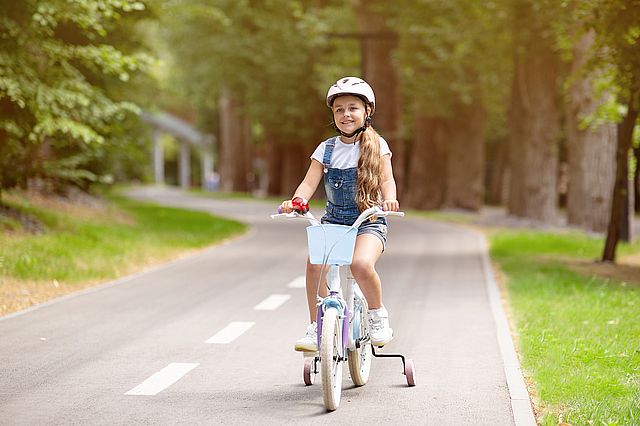 Zegarek GPS dla dziecka u dziewczynki jadącej na rowerze po parku ubranej w białą bluzkę, ogrodniczki i biały kask